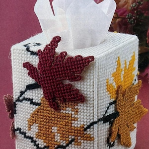May include: A white tissue box cover with a crocheted design featuring fall leaves in shades of red, yellow, and black.
