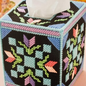 May include: A black tissue box cover with a colourful geometric pattern featuring pink, blue, green, and purple flowers. The cover is made of yarn and has a square shape.