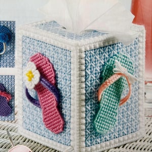 May include: A blue, green, and pink tissue box cover with a white border and a woven pattern. The cover features three embroidered flip-flops with flowers.