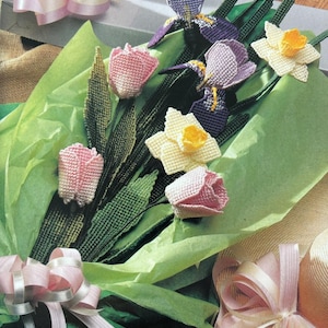 May include: A bouquet of crocheted flowers in shades of pink, purple, yellow, and green. The flowers are arranged in green tissue paper wrapping with a pink ribbon.