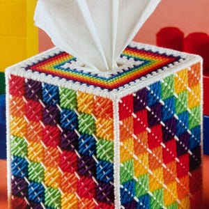 May include: A square tissue box cover with a vibrant, multicolored design. The cover is crafted with a cross-stitch pattern in a rainbow of colors, including red, orange, yellow, green, blue, and purple. White tissues are visible at the top.