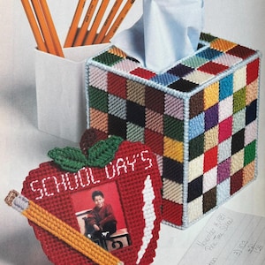 Puede incluir: Una funda para caja de pañuelos de lona plástica con un patrón de tablero de ajedrez multicolor y un marco de fotos rojo en forma de manzana con el texto SCHOOL DAY'S, un diseño de lápiz amarillo y la foto de un niño, junto a varios lápices.