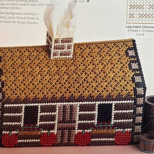 May include: A handcrafted tissue box cover shaped like a log cabin. The roof is a woven pattern in shades of brown and gold. The cabin features windows, a chimney, and red accents. Two small wooden barrels are next to the cabin.