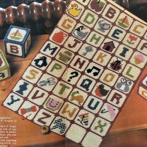 K&ouml;nnte beinhalten: Ein handgefertigtes Alphabet-Muster mit gestickten Quadraten, die Buchstaben und verschiedene Bilder wie eine Ente, einen Apfel und eine Eule zeigen. Drei dekorative Bl&ouml;cke mit gestickten Designs sind ebenfalls vorhanden. Das Muster wird auf einer Holzoberfl&auml;che pr&auml;sentiert.