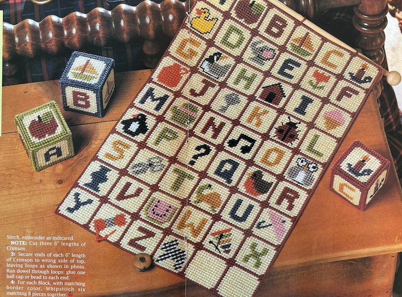 K&ouml;nnte beinhalten: Ein handgefertigtes Alphabet-Muster mit gestickten Quadraten, die Buchstaben und verschiedene Bilder wie eine Ente, einen Apfel und eine Eule zeigen. Drei dekorative Bl&ouml;cke mit gestickten Designs sind ebenfalls vorhanden. Das Muster wird auf einer Holzoberfl&auml;che pr&auml;sentiert.