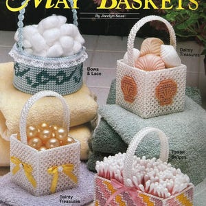 Könnte beinhalten: Vier Körbe aus Kunststoffgewebe mit dem Titel May Baskets von Jocelyn Sass. Ein runder Korb mit weißer Spitze und Wattebällchen, ein quadratischer Korb mit Muschelmotiven, ein quadratischer Korb mit gelben Schleifen und Perlen sowie ein gestreifter Korb.