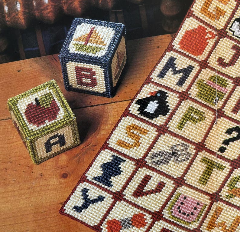 K&ouml;nnte beinhalten: Handgefertigte Kreuzstich-Alphabetbl&ouml;cke mit Segelboot-, Apfel- und Buchstabenmotiven. Die Bl&ouml;cke sind in Gr&uuml;n-, Blau- und Rott&ouml;nen gehalten. Ein passendes Kreuzstich-Alphabetmuster ist ebenfalls sichtbar.