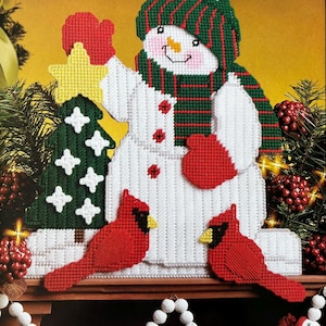 May include: A plastic canvas craft project featuring a white snowman with a green and red striped hat and scarf, red mittens, and three red buttons. The snowman holds a yellow star atop a green Christmas tree, with two red cardinal birds below.