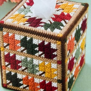 May include: A square tissue box cover with a patchwork design of fall leaves in shades of red, orange, yellow, green, and brown. The cover is made of yarn and has a brown border.