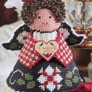 May include: A handmade felt angel ornament with a red heart that says "Love Me" on it. The angel has brown curly hair, a red and white checkered dress, and a black skirt with green and white floral designs.