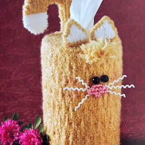 May include: A crocheted orange cat tissue box cover with white accents. The cat has black eyes, a pink nose, and white whiskers. The tissue box is visible inside the cover.