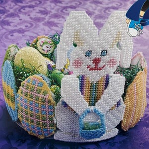 May include: A handcrafted Easter basket featuring a white bunny holding a blue basket. The basket is filled with colorful, decorated eggs and green faux grass. The bunny has blue eyes, pink cheeks, and a rainbow striped vest.