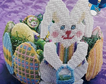 May include: A handcrafted Easter basket featuring a white bunny holding a blue basket. The basket is filled with colorful, decorated eggs and green faux grass. The bunny has blue eyes, pink cheeks, and a rainbow striped vest.