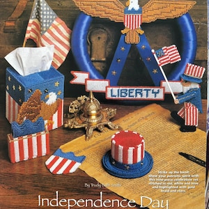May include: A collection of patriotic Independence Day decorations, including a tissue box, a wreath with an eagle, and miniature flags. The items are primarily red, white, and blue, with gold accents. The word "LIBERTY" is displayed on a banner.