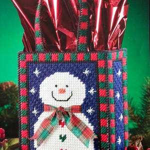 May include: A blue and white canvas tote bag with a snowman design. The bag has a red and green tartan ribbon around the snowman's neck and a red and green tartan border. The bag has a red and green tartan handle.