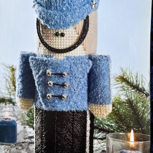 May include: A blue and white felt Christmas nutcracker ornament with silver buttons and stars. The nutcracker is wearing a blue hat and has a black and white striped skirt.
