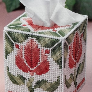 May include: A square tissue box cover with a needlepoint design. The cover features a white background with red tulip and green leaf patterns. White tissues are emerging from the top opening. The design is a classic, handmade style.