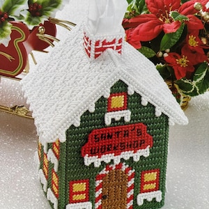 Puede incluir: Un cubretisú navideño artesanal con forma de taller de Santa. La casa verde presenta un tejado blanco cubierto de nieve, detalles rojos y amarillos, y el texto "SANTA'S WORKSHOP". Una chimenea y pañuelos sobresalen.