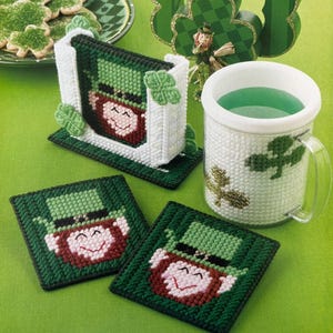 May include: A St. Patrick's Day themed set featuring a mug, coasters, and a napkin holder, all adorned with leprechaun faces and shamrock designs. The mug is filled with green liquid. The items are made of a woven material.