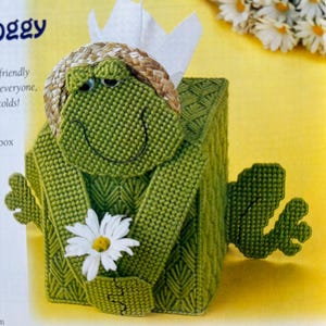 May include: A green crocheted frog tissue box cover with a straw hat and a white daisy. The frog is smiling and has a friendly expression. The text "Boggy" is visible in the image.