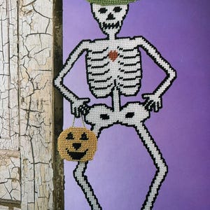 May include: A Halloween-themed decoration featuring a skeleton figure made of white and black yarn. The skeleton wears a green hat and holds a small pumpkin-shaped bag. The background is a solid purple color.