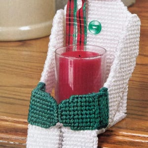 May include: A plastic canvas snowman candle holder featuring a white body, a black top hat with a green band, and green mittens. The snowman wears a red and green plaid scarf, has a green button on its chest, and holds a red glass candle votive.
