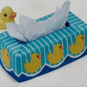 May include: A rectangular tissue box cover with a blue and white striped pattern and yellow rubber ducky illustrations. A yellow rubber duck sits next to the tissue box. White tissues are coming out of the top.