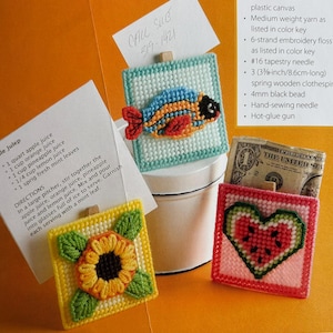 May include: Four colorful plastic canvas crafts with wooden clothespins. The crafts are a fish, a sunflower, a watermelon heart, and a recipe card holder. The recipe card holder has the text "CALL SUE 319-1421" on it.