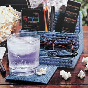 May include: A blue plastic yarn craft organizer with multiple compartments. The organizer has a remote control, pens, pencils, and glasses inside. There is a glass of water and popcorn on a blue coaster.