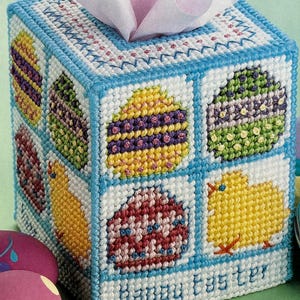 May include: A white tissue box cover with a blue border and a colorful Easter egg design. The cover has a stitched design of eggs, chicks, and the words "Happy Easter".