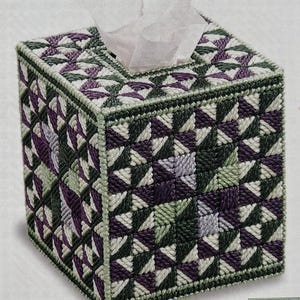 May include: A square tissue box cover with a geometric pattern in shades of purple, green, and white. The cover is made of a woven material, with a decorative border. White tissues are visible at the top.