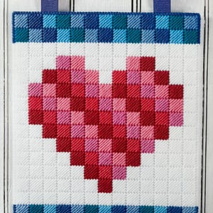 Op de afbeelding: Een wit canvas met een rood, roze en wit gepixelde hartontwerp. Het canvas heeft een blauwe en turquoise geruite rand.