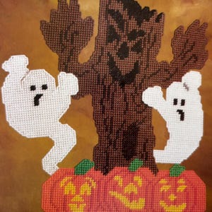 May include: A Halloween needlepoint design. It features a brown tree with a smiling face and outstretched arms, two white ghosts, and three orange pumpkins with green stalks. The background is a warm, textured brown.