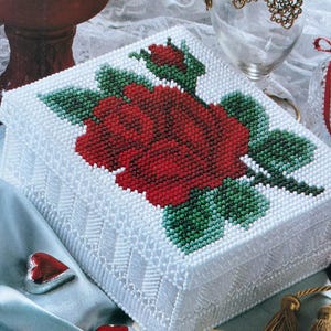 Może przedstawiać: Białe pudełko z koralików z wzorem czerwonej róży i zielonych liści. Pudełko jest ozdobione wzorem z białych koralików wokół krawędzi.