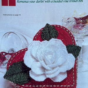 May include: A red heart-shaped beaded trinket box with a white rose and green leaves. The box is decorated with silver beads around the edges. The text "Heart of the Rose" and "Romance your darlin' with a beaded rose trinket box" is printed on the page. Instructions on page 18.