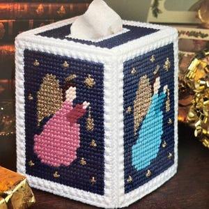 May include: A white tissue box cover with a blue background and two angels with gold wings. The angels are wearing pink and blue dresses and are surrounded by gold stars.