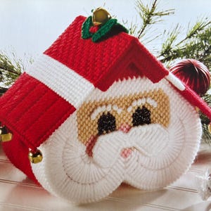 May include: A white and red crocheted Santa Claus tissue box cover with a red roof and gold bells. The Santa Claus face is white with a pink nose and black eyes.
