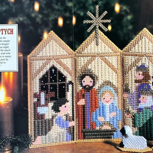 May include: A plastic canvas nativity triptych featuring three hinged panels with stitched figures of the Holy Family and wise men, topped with a gold star. A sign reads NATIVITY TRIPTYCH. Two small white sheep figures stand in front of the panels.