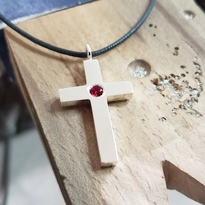 Puede incluir: Un colgante de cruz de plata con una piedra preciosa roja en el centro, colgado de un cordón negro.