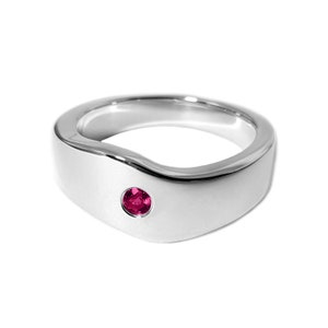 May include: A silver ring with a smooth, curved design. A small, round, bright pink gemstone is set into the side of the ring. The ring has a modern, minimalist style. The ring is set against a white background.