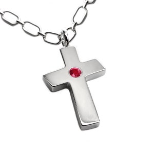 Puede incluir: Un colgante de cruz de plata con una pequeña piedra preciosa roja en el centro, colgando de una cadena de plata.