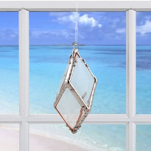 May include: A clear glass ornament with a silver frame, hanging from a white string in front of a window with a view of the ocean and beach.
