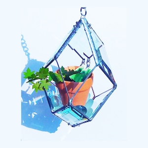 May include: A hanging glass terrarium with a geometric shape, featuring a small potted plant and blue glass stones. The terrarium is made of clear glass with silver accents and hangs from a silver chain.