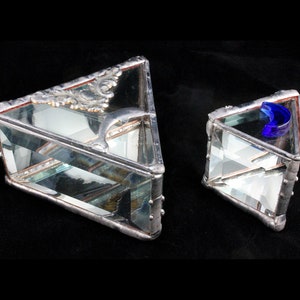 May include: Two triangular glass boxes with silver trim. The larger box has a decorative silver piece on top and a blue glass dolphin inside. The smaller box has a blue glass crescent moon inside.