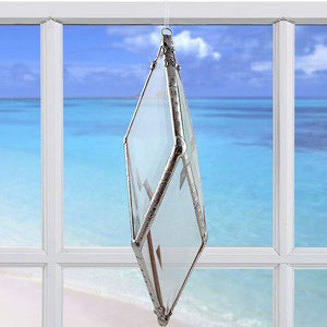May include: A clear glass diamond-shaped suncatcher with silver accents hanging in a window. The suncatcher is reflecting light from the window.