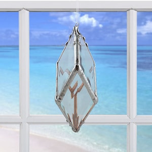 May include: A silver and clear glass suncatcher with a geometric design. The suncatcher is hanging in a window with a view of the ocean.
