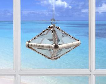 May include: A clear glass diamond-shaped ornament with silver accents hanging in a window with a view of a beach and ocean.