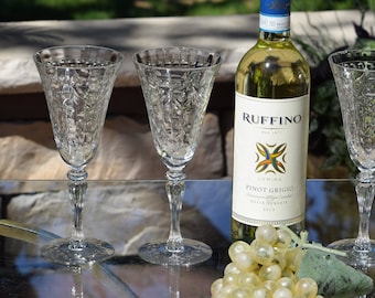 Może przedstawiać: Cztery przezroczyste, grawerowane kieliszki do wina stoją na szklanym stole z butelką wina Pinot Grigio Ruffino i gronem zielonych winogron.