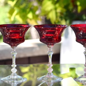 May include: Three ruby red glass cocktail coupes with wide bowls and clear, textured stems, positioned next to a tall, rectangular glass bottle labelled North Shore Distillery Distiller's Gin No. 6 with a martini glass graphic and text.