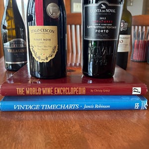 May include: A collection of wine bottles on a wooden table, including a bottle of Pinot Noir, a bottle of Port, and a bottle of white wine. Two books, "The World Wine Encyclopedia" and "Vintage Timecharts", are stacked on the table.